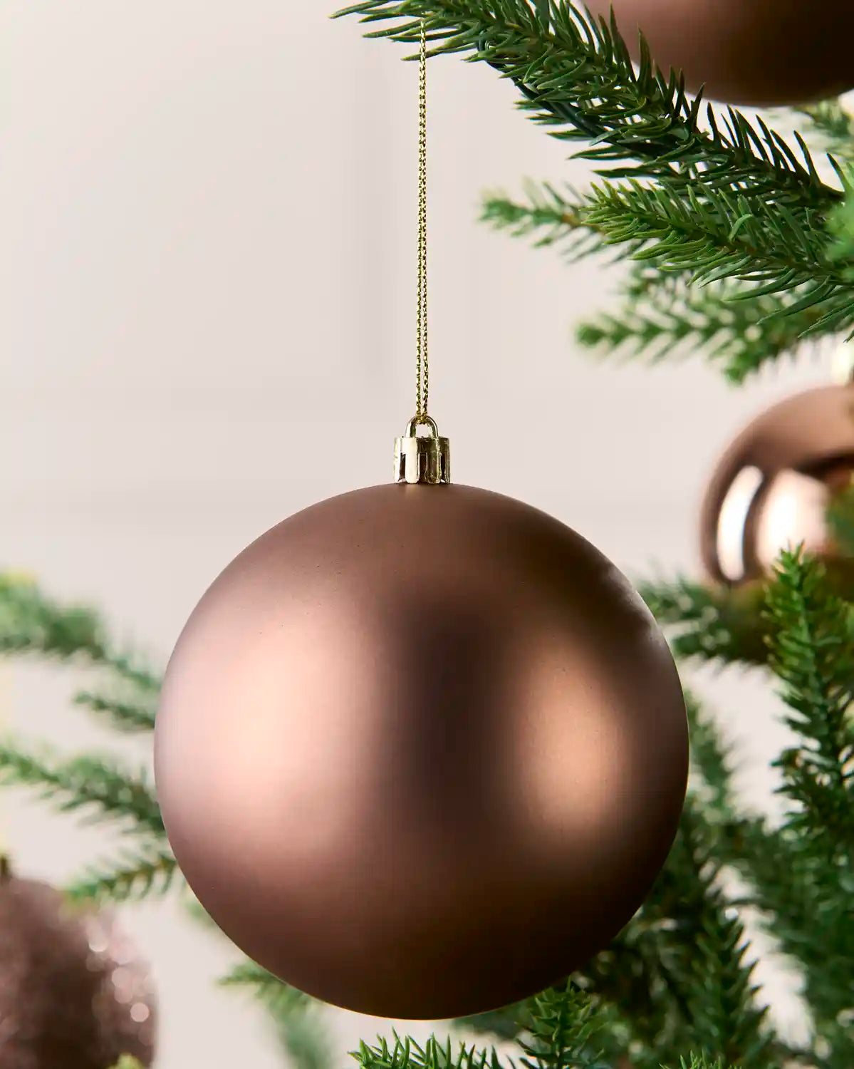 A matte mocha Christmas bauble ornament hanging from a gold string on a green Christmas tree branch.