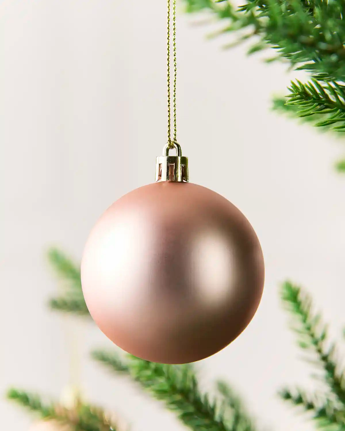 A matte rose gold Christmas bauble hanging from a gold string against a soft white background with blurred green pine branches.