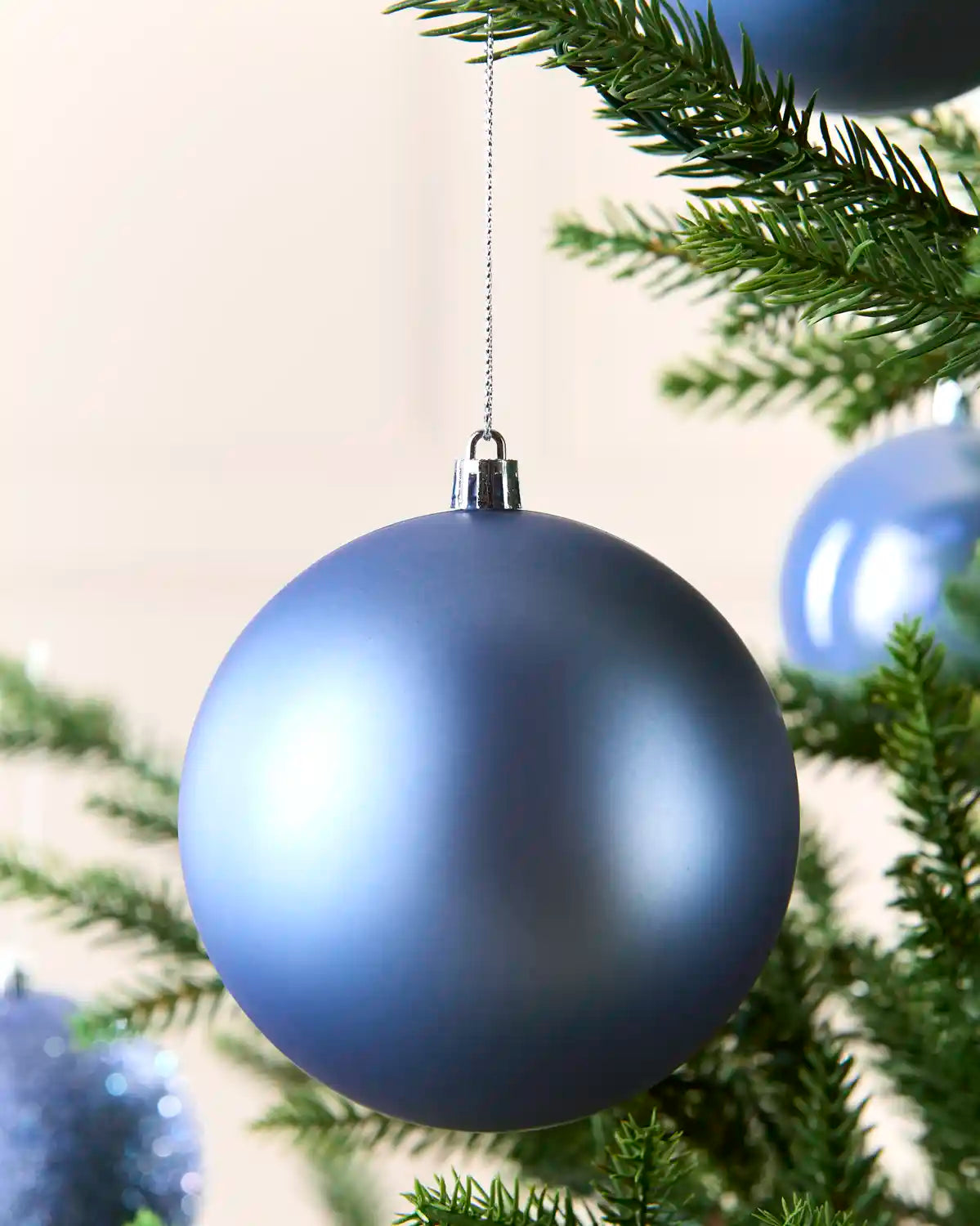 A matte slate blue Christmas tree ornament hanging from a silver cap on a silver string, nestled among green pine tree branches.