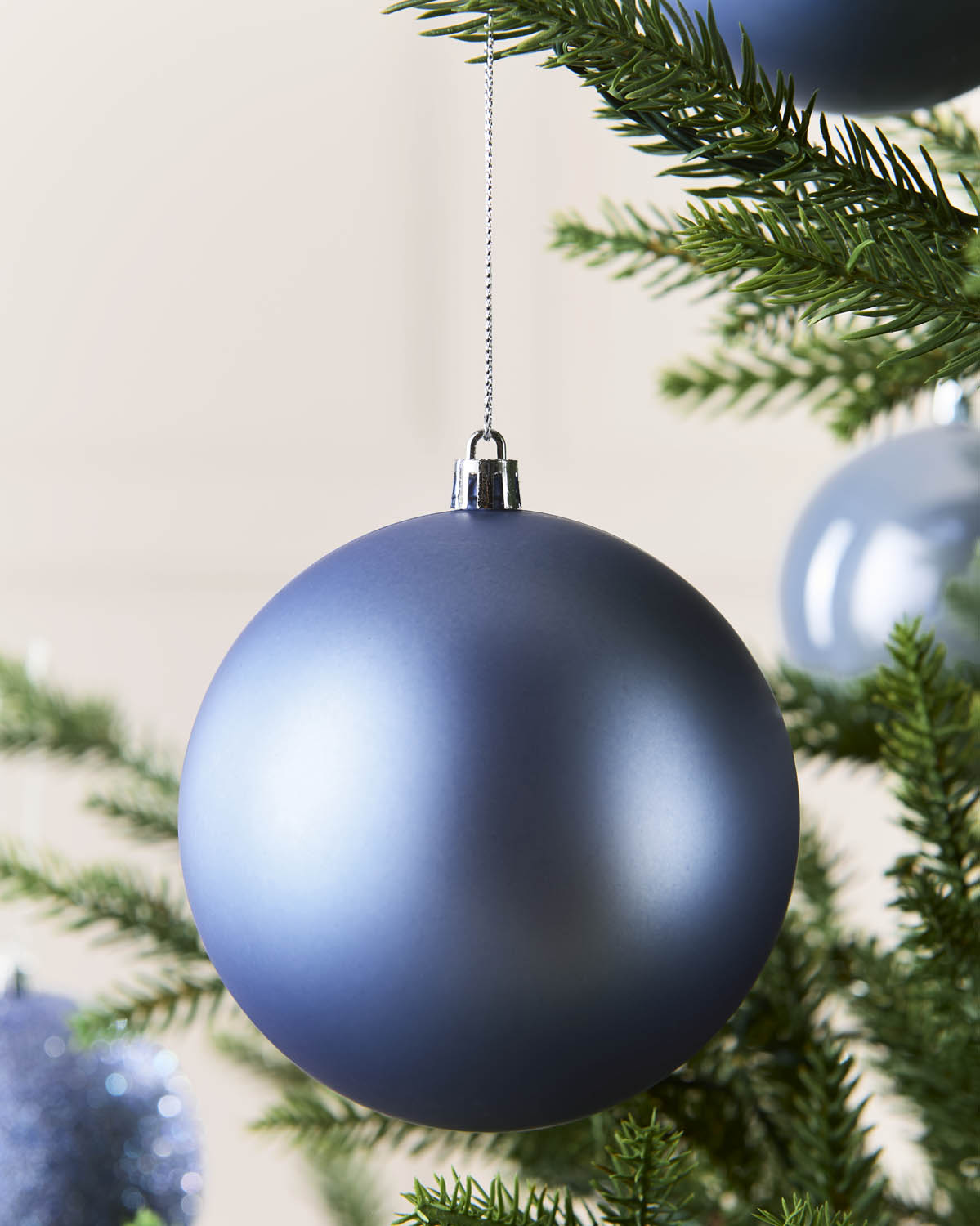 A matte slate ice blue Christmas tree ornament hanging from a silver cap on a silver string, nestled among green pine tree branches.
