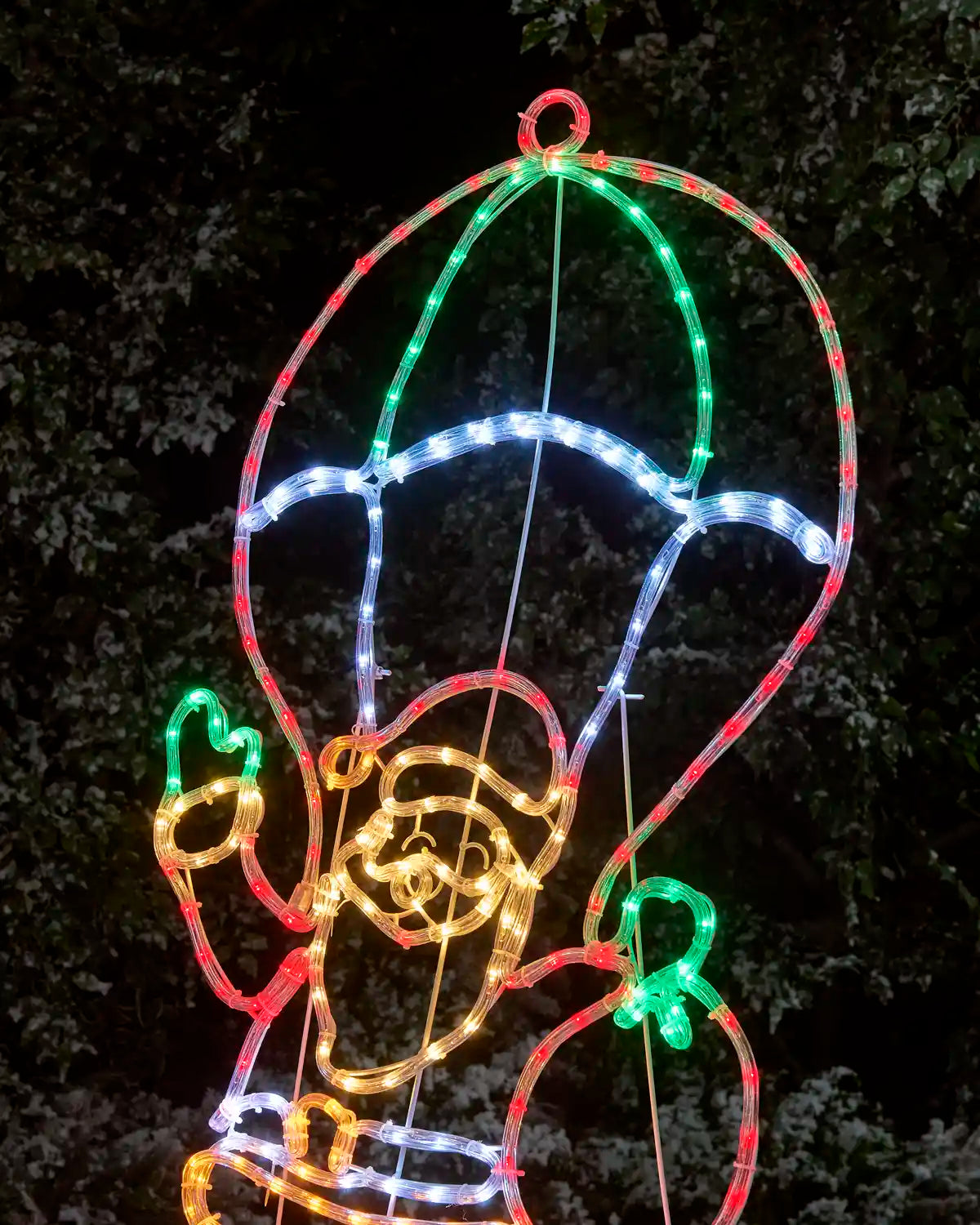 A multicolor LED light Christmas decoration shaped like Santa Claus with a hot air balloon, photographed outdoors at night against a dark, leafy background.