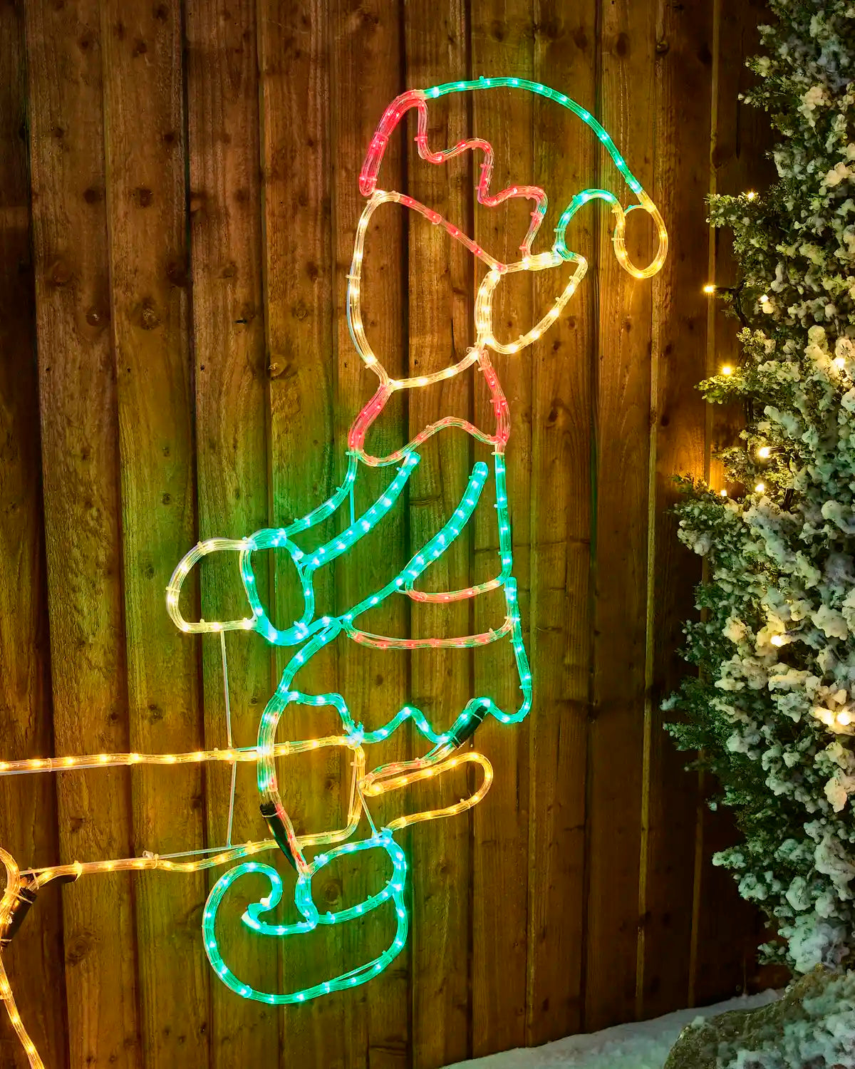 A multicolour LED light Christmas decoration shaped like an elf figure, mounted against a dark wooden fence with a snowy green tree on the right side.