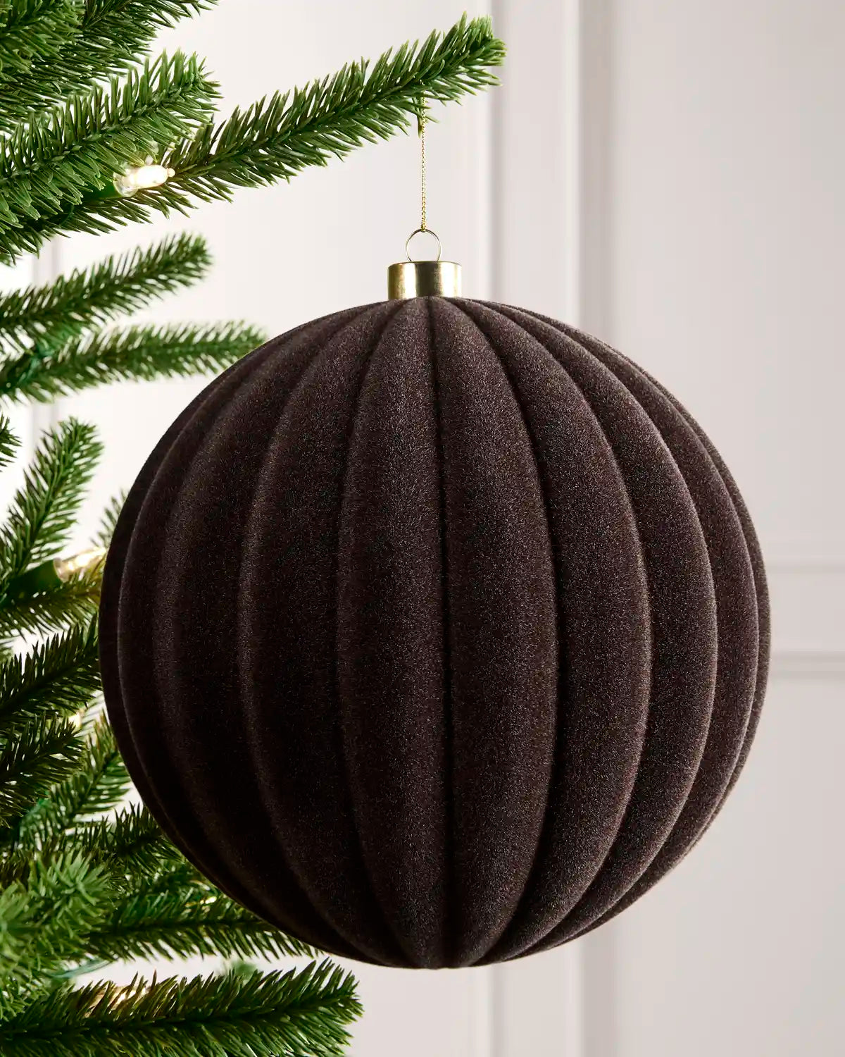 A n espresso coloured ribbed fabric Christmas ornament hanging from a green pine tree branch against a white paneled wall.