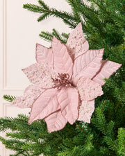 A pale pink artificial poinsettia flower with textured, pearl leaves, displayed on a green pine Christmas tree against a white panelled wall.