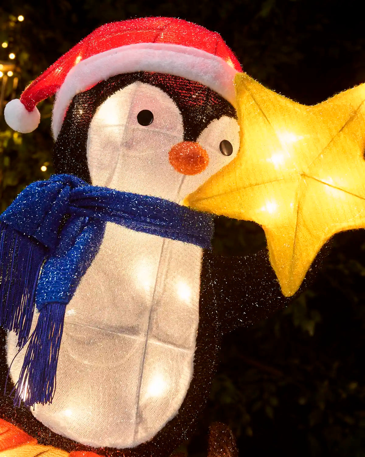 A penguin-shaped holiday decoration with a red Santa hat, blue scarf, and a glowing yellow star beside it, photographed outdoors at night against a dark background.