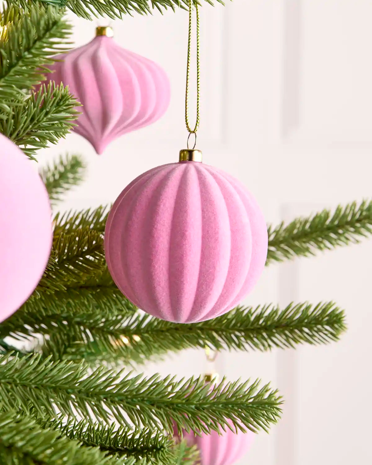 A pink ribbed velvet Christmas ornament hanging from a green pine tree branch, with other matching pink ornaments partially visible in the background.