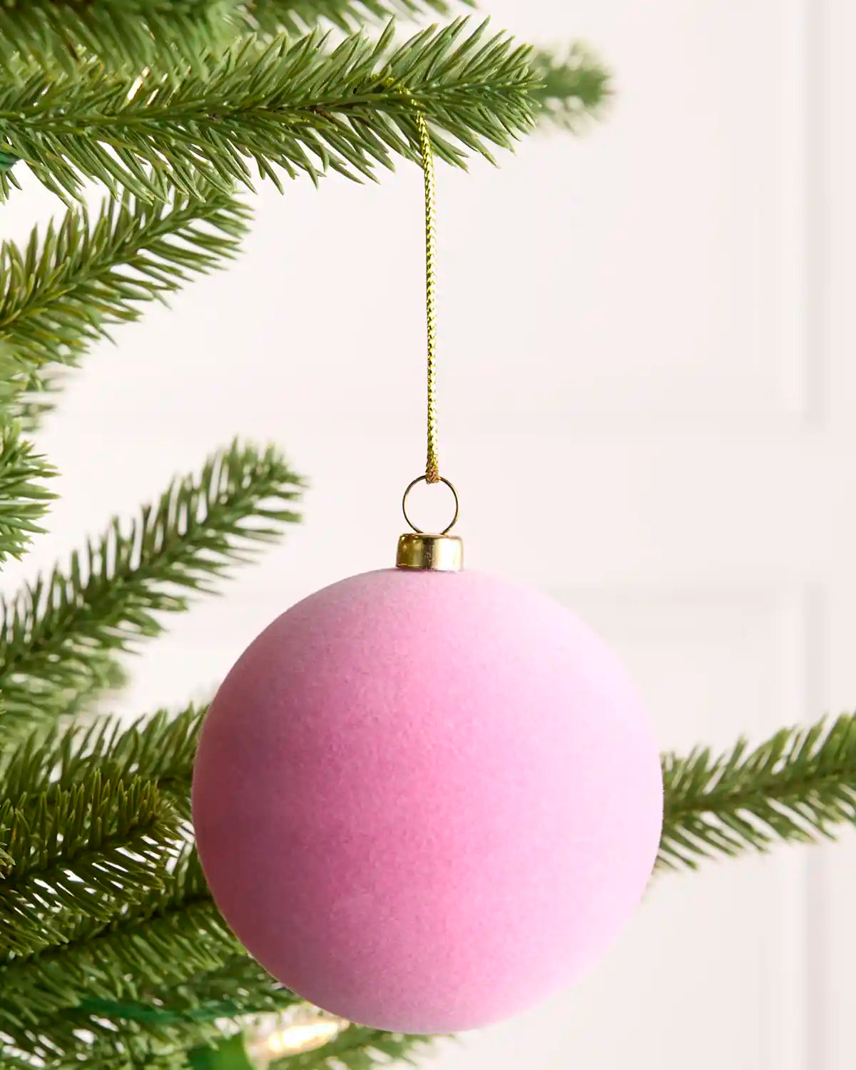 A pink velvet Christmas ornament hanging from a gold string on a green pine tree branch against a soft white background.