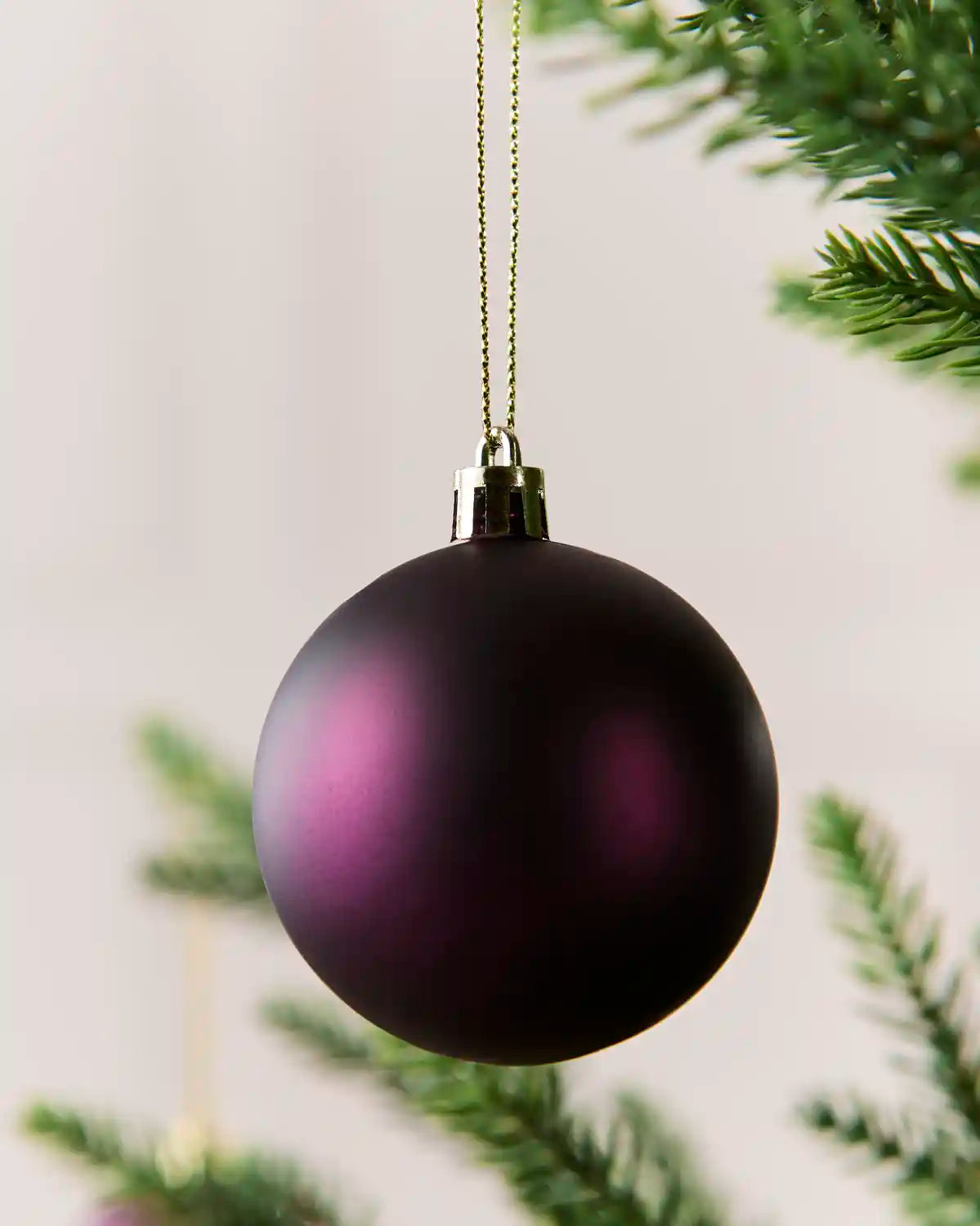 A purple matte Christmas bauble hanging from a thin gold string, with blurred green pine branches in the background.