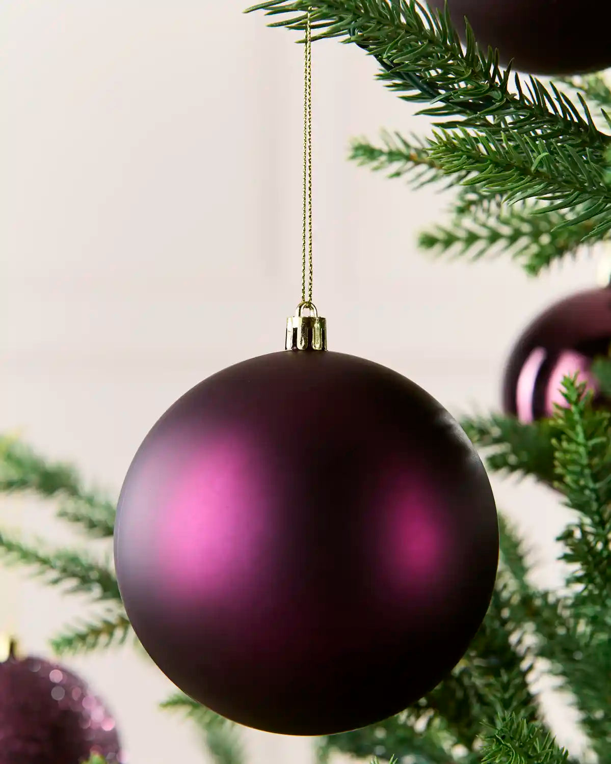 A purple matte Christmas bauble ornament hanging from a gold string on a green Christmas tree branch.