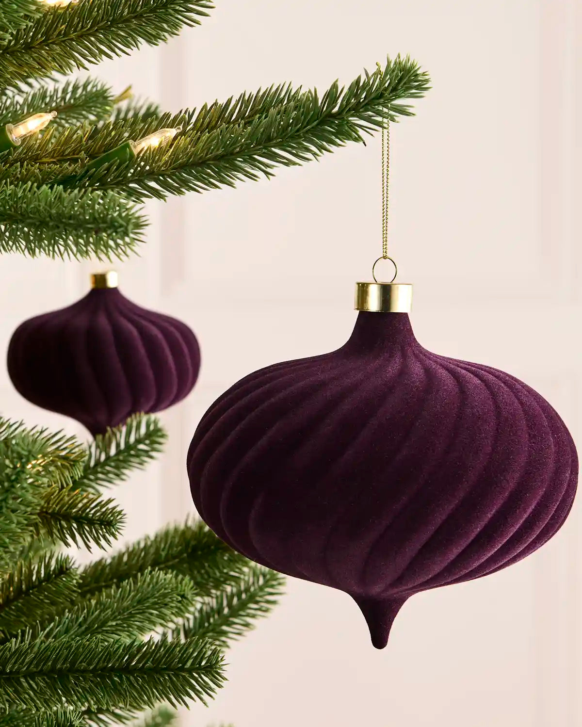A purple ribbed velvet Christmas ornament hanging from a gold cap on a gold string, displayed next to green pine tree branches with warm string lights against a soft cream background.