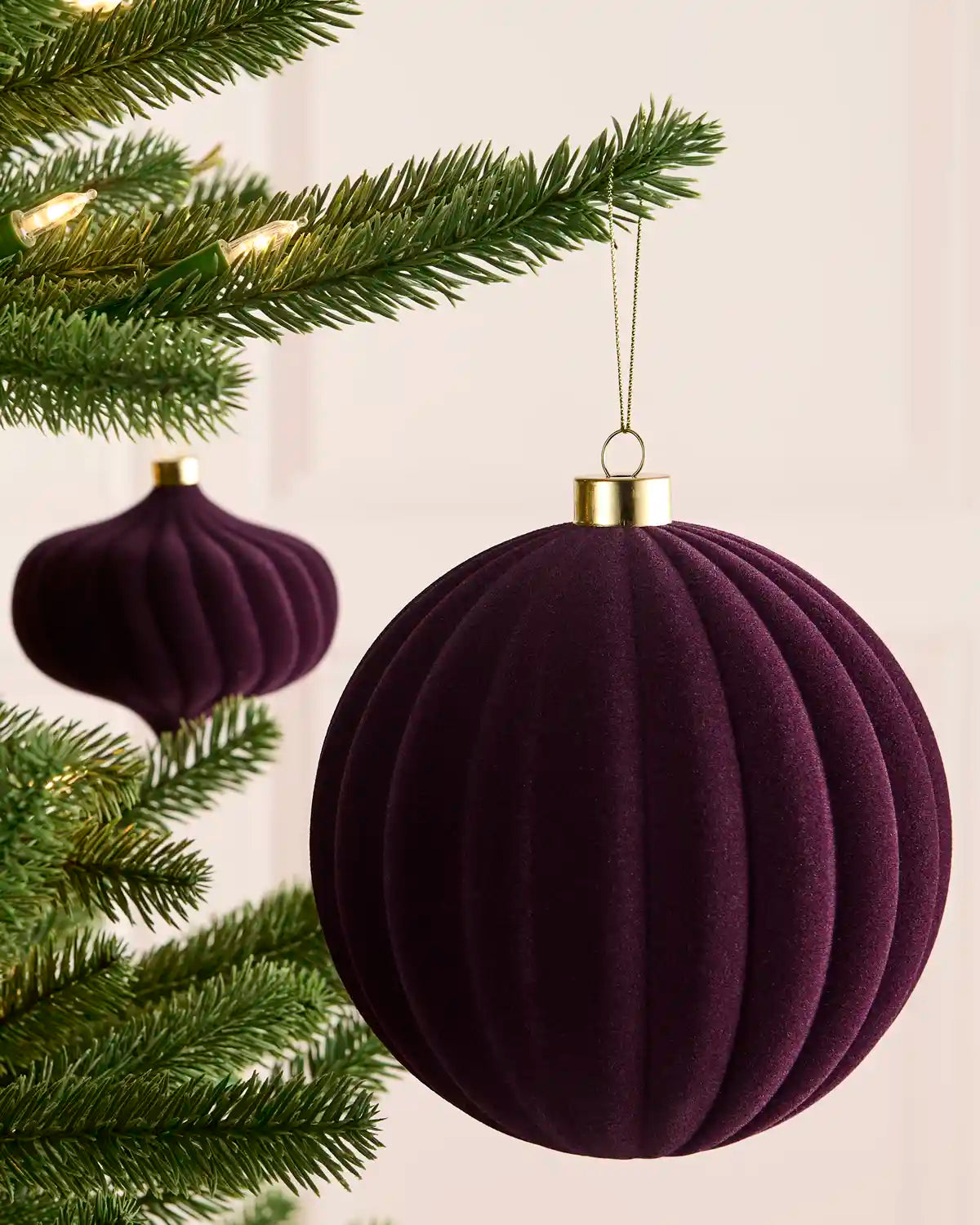A purple ribbed velvet Christmas ornament hanging from a green Christmas tree branch, with a gold cap and a thin gold hanger. A second matching ornament is visible in the background.