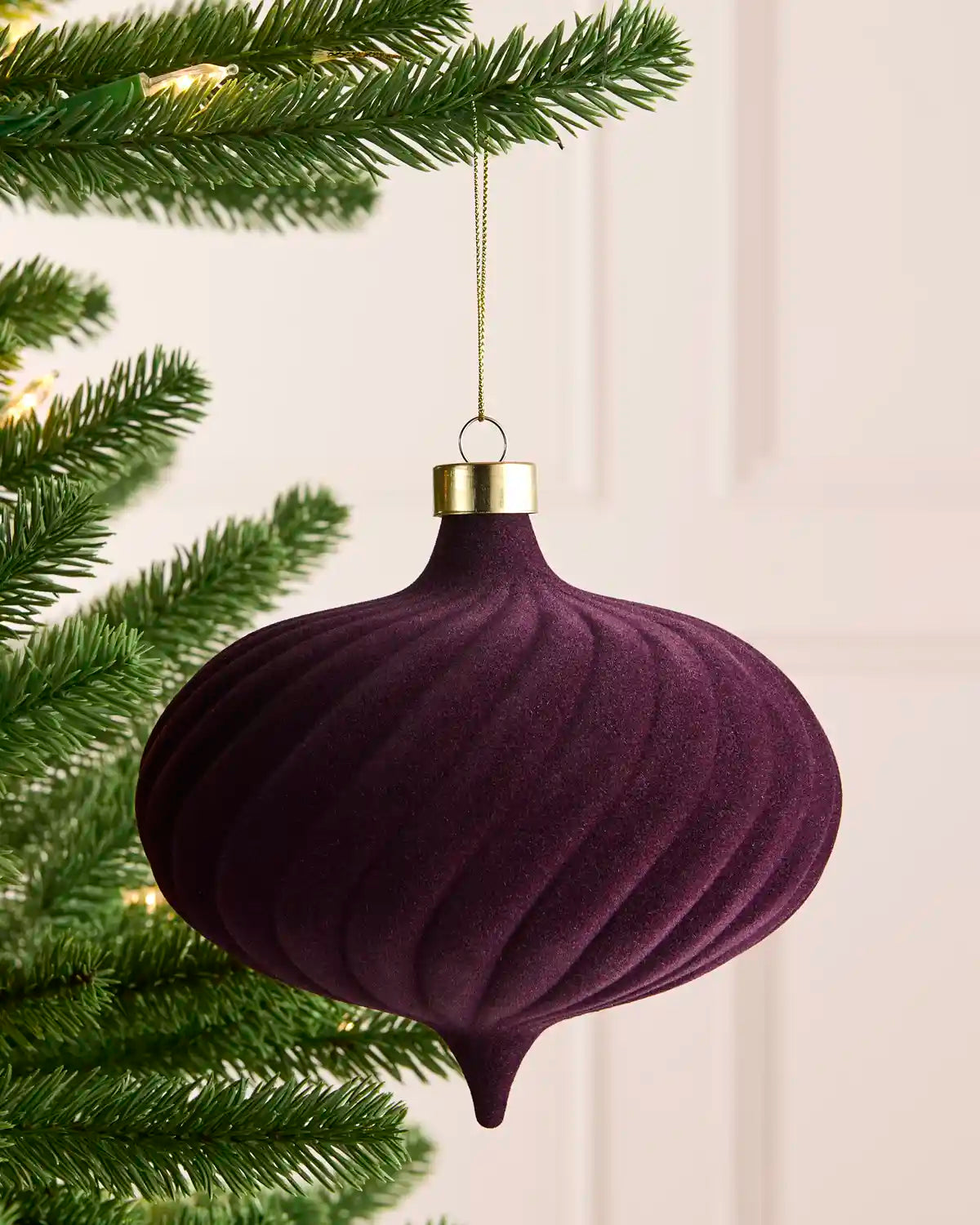 A purple ribbed velvet Christmas ornament hanging from a green pine tree branch with warm string lights in the background.