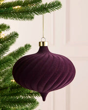 A purple ribbed velvet Christmas ornament hanging from a green pine tree branch with warm string lights in the background.