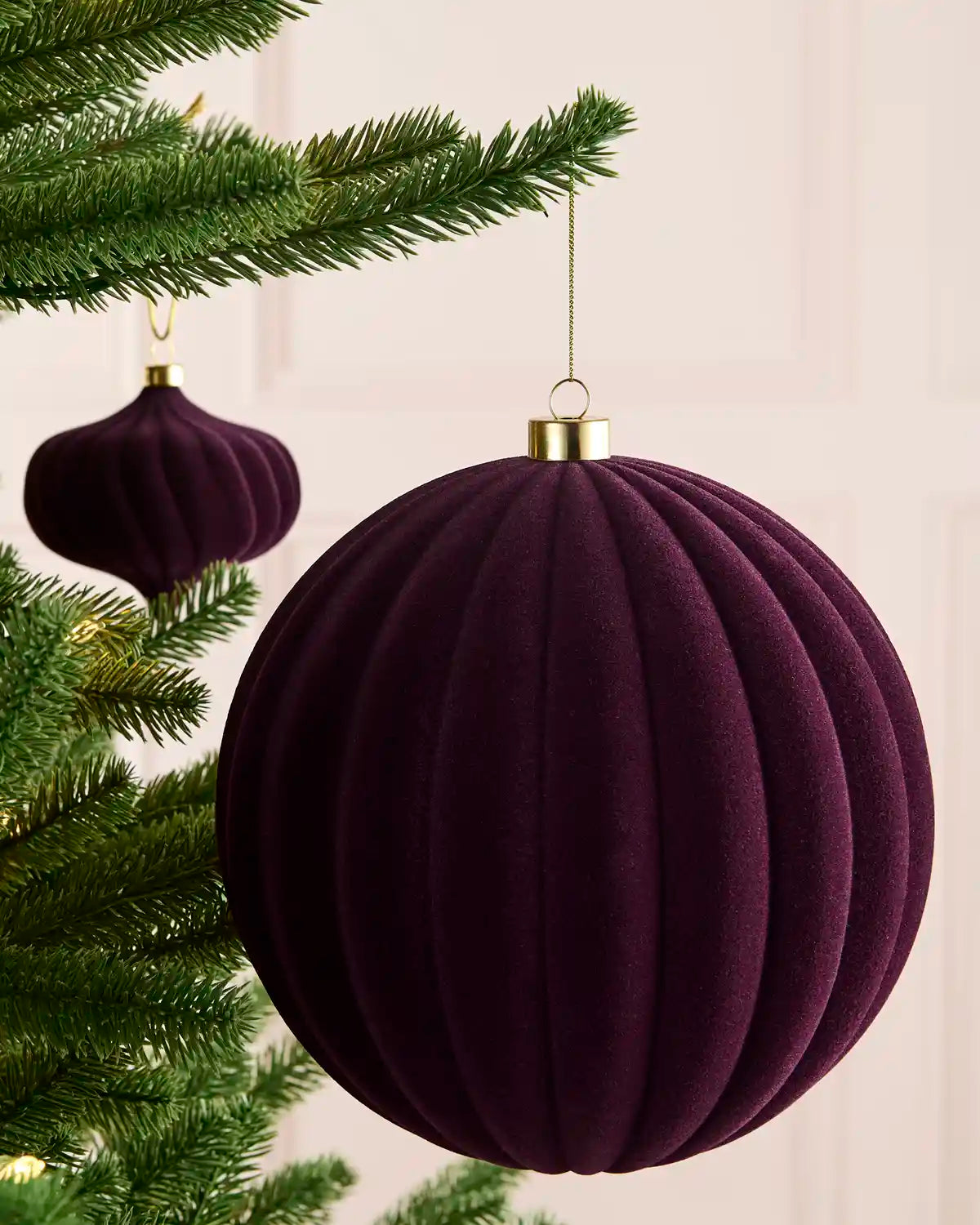 A purple ribbed velvet Christmas tree ornament hanging from a green pine tree branch, with a gold cap and a thin hanging cord. A second matching ornament is visible in the background.