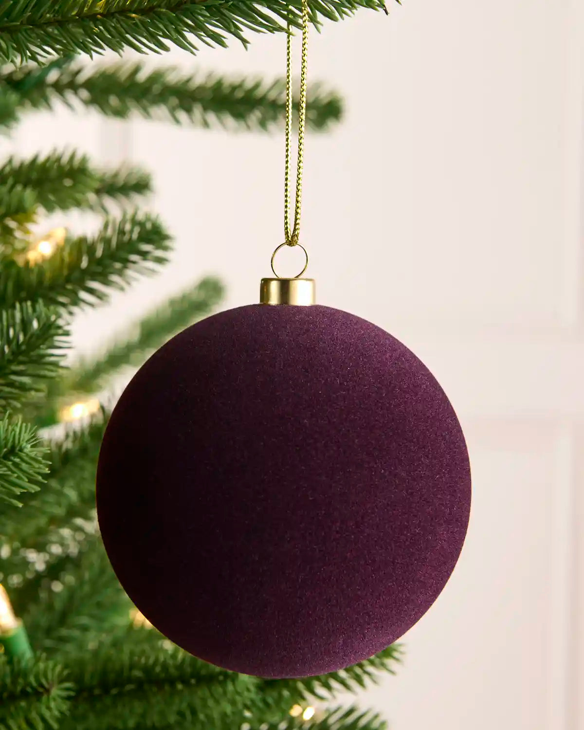 A purple velvet Christmas bauble ornament hanging from a gold string on a green Christmas tree branch.