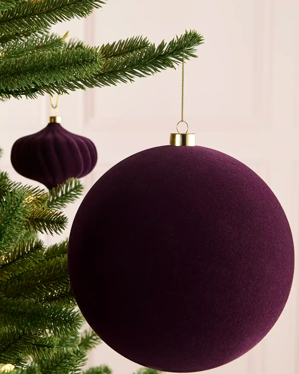 A purple velvet Christmas bauble ornament hanging from a green Christmas tree branch, with a smaller matching ornament in the background.