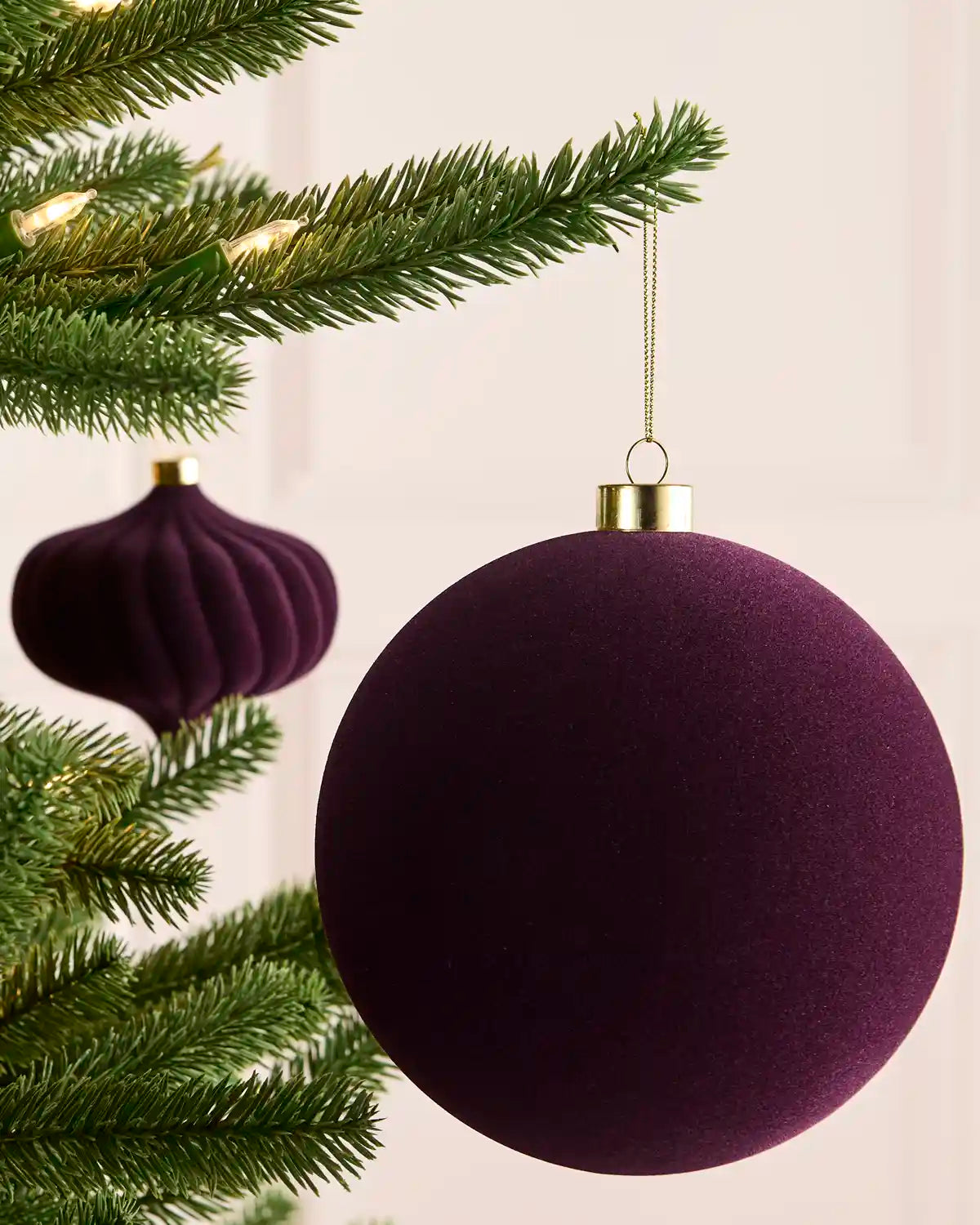 A purple velvet Christmas bauble ornament hanging from a green Christmas tree branch with warm string lights, with a second matching ornament blurred in the background.
