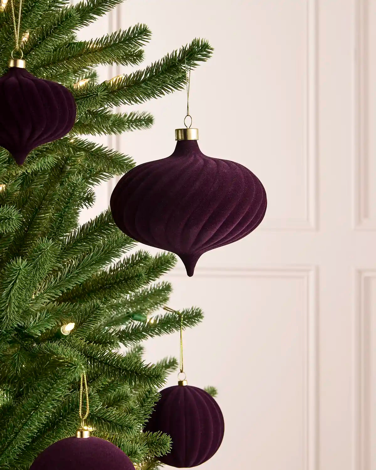 A purple velvet Christmas tree ornaments hanging on a green pine tree, with a soft cream panelled wall in the background.