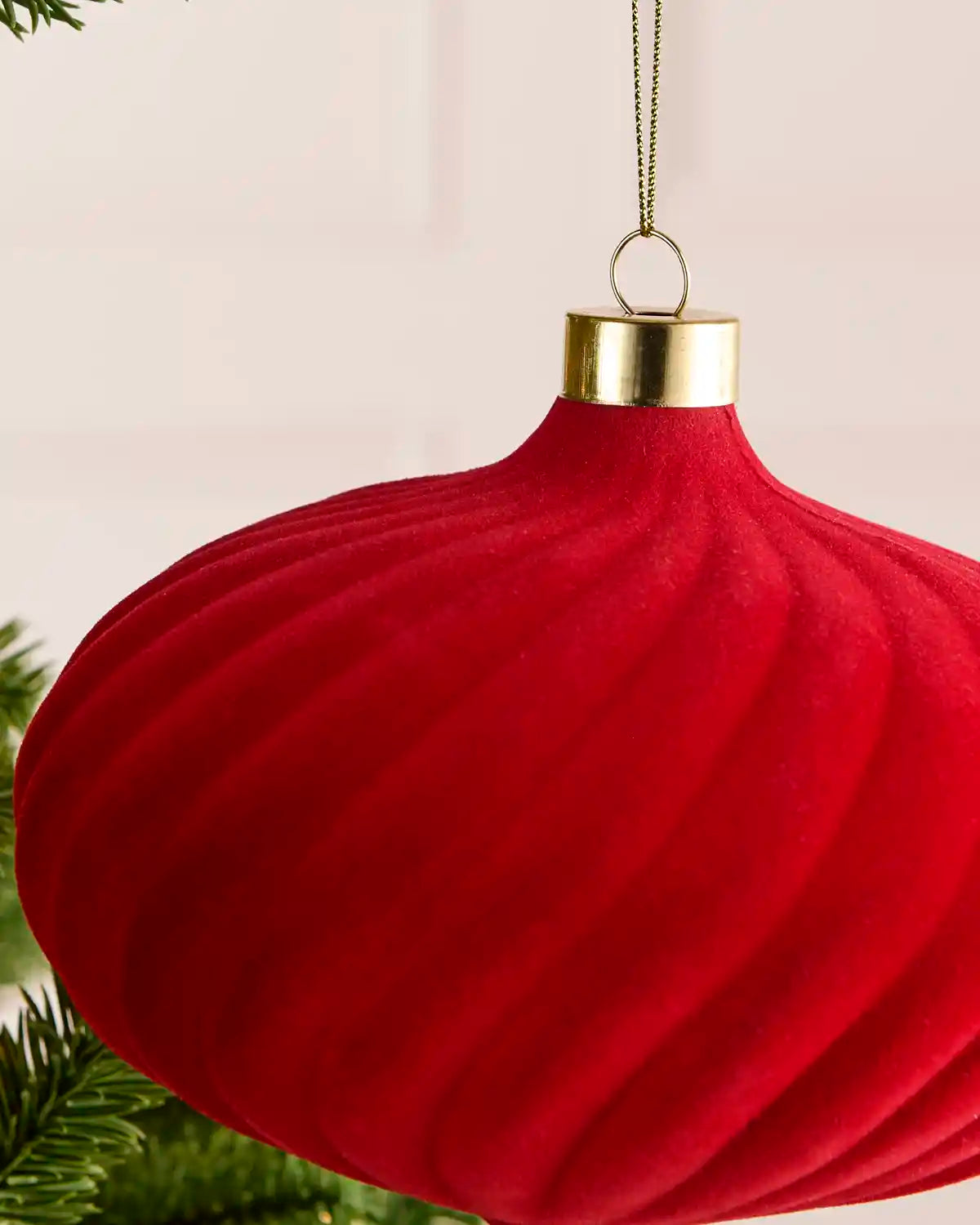 A red ribbed Christmas ornament hanging from a gold cap with a gold hanging loop, shown against a soft light background with a hint of green pine branches at the bottom left.