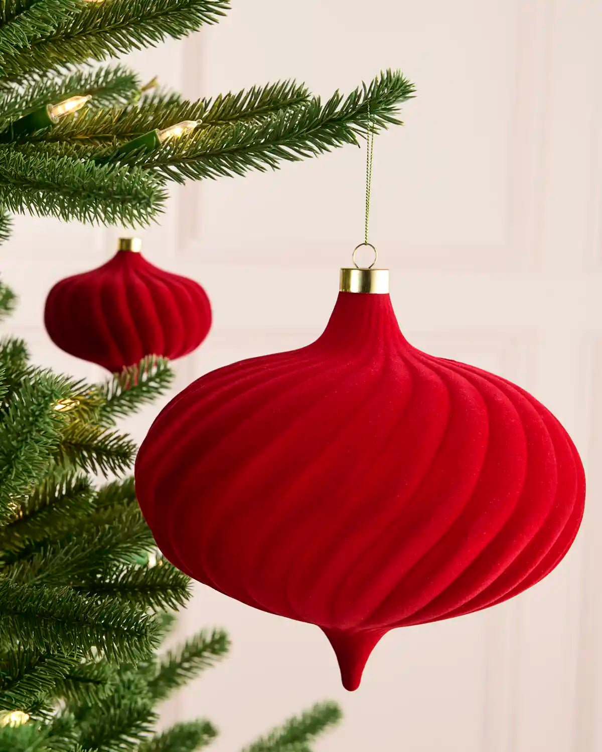 A red ribbed Christmas ornament hanging from a green Christmas tree branch, with a second red ornament blurred in the background.