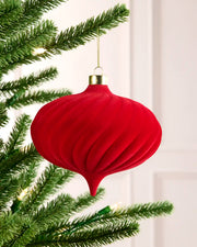 A red ribbed Christmas tree ornament hanging from a green pine tree branch with warm white string lights in the background.