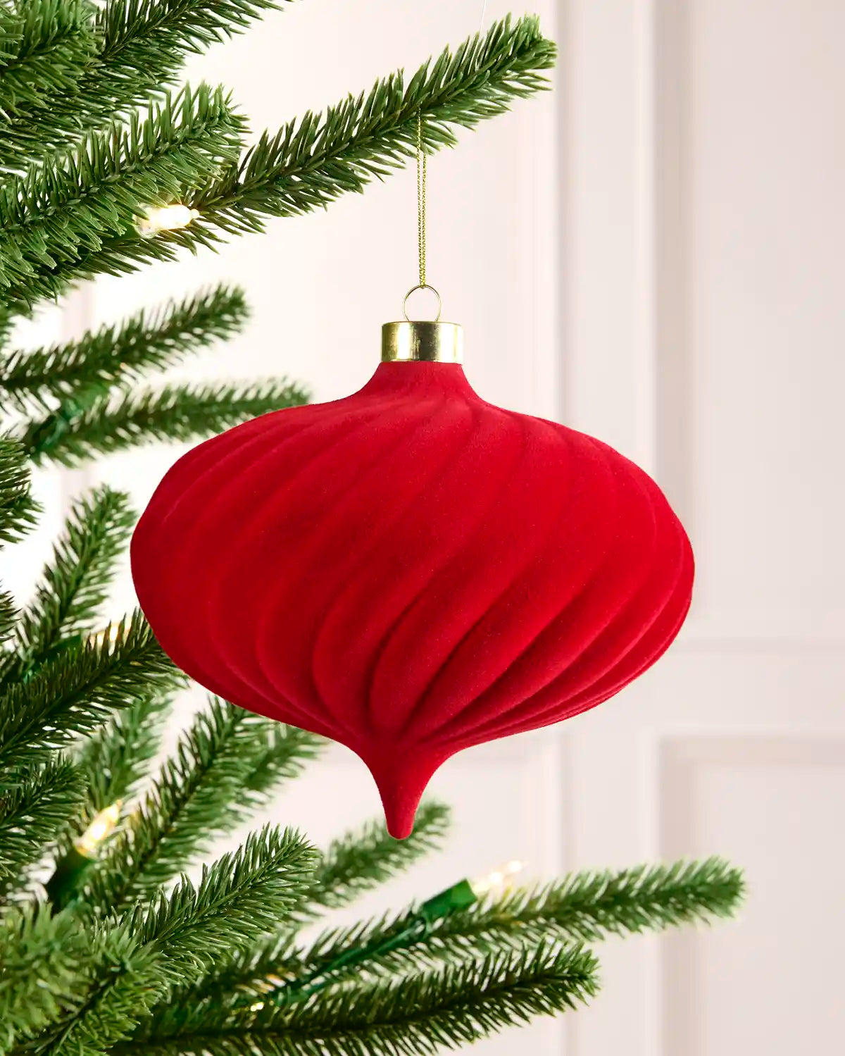 A red ribbed Christmas tree ornament hanging from a green pine tree branch with warm white string lights in the background.