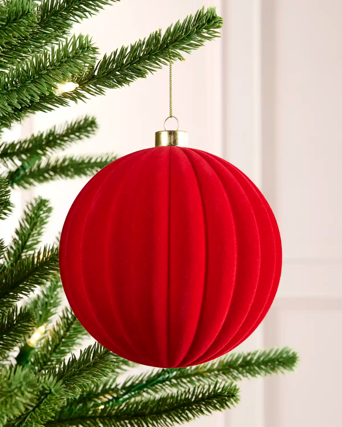 A red ribbed fabric Christmas ornament hanging from a green Christmas tree branch with warm string lights in the background.