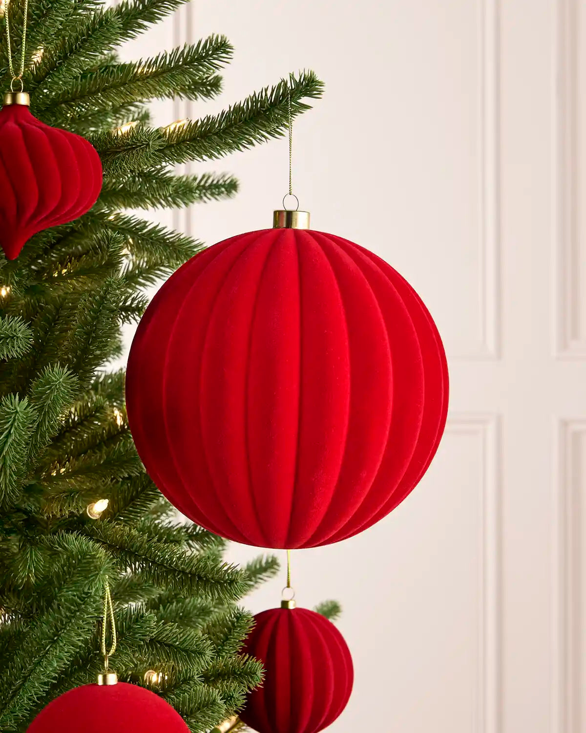 A red ribbed fabric Christmas ornament hanging from a green Christmas tree with warm white string lights, against a light cream paneled wall.
