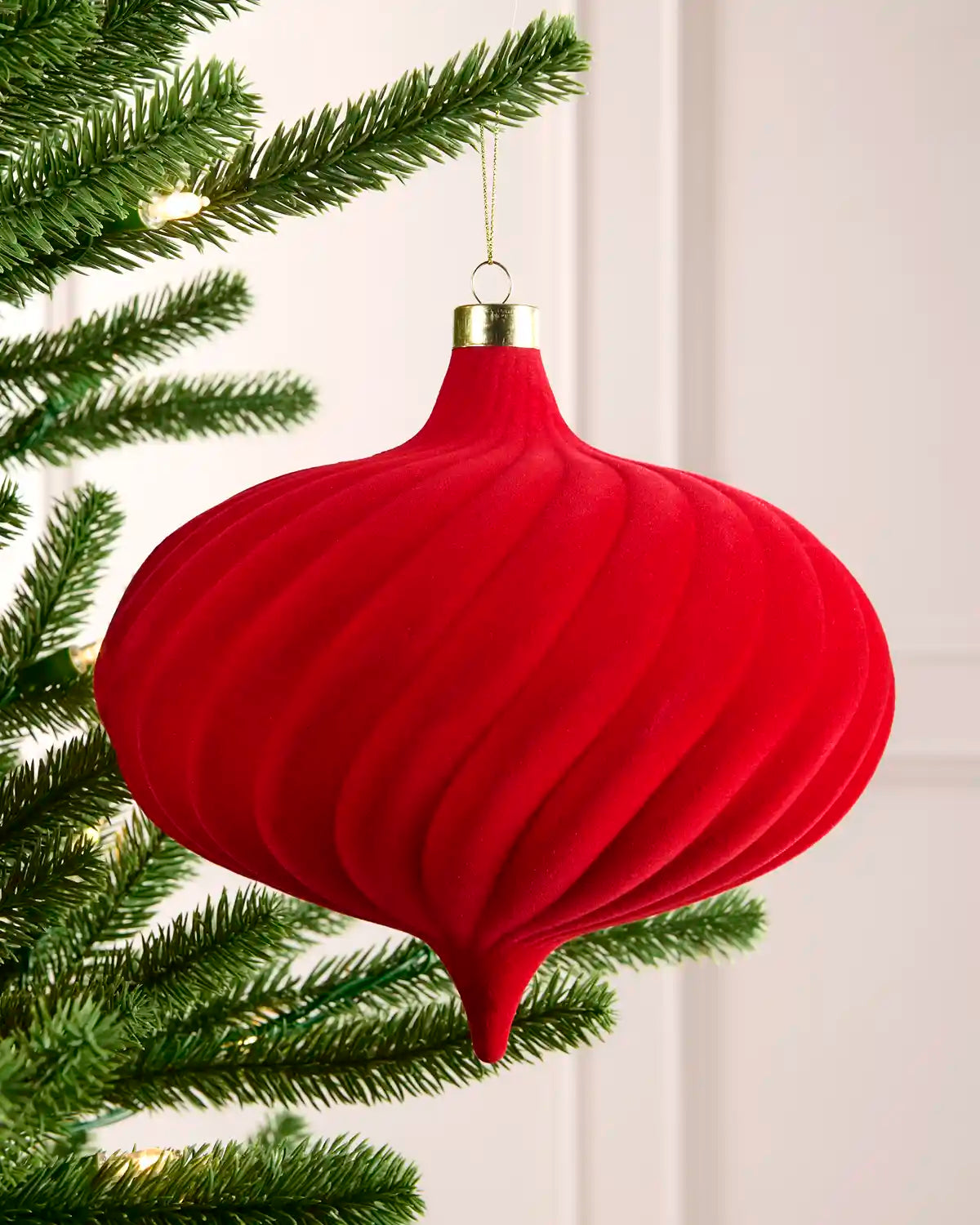 A red ribbed fabric Christmas tree ornament hanging from a green pine tree branch against a soft off-white paneled background.