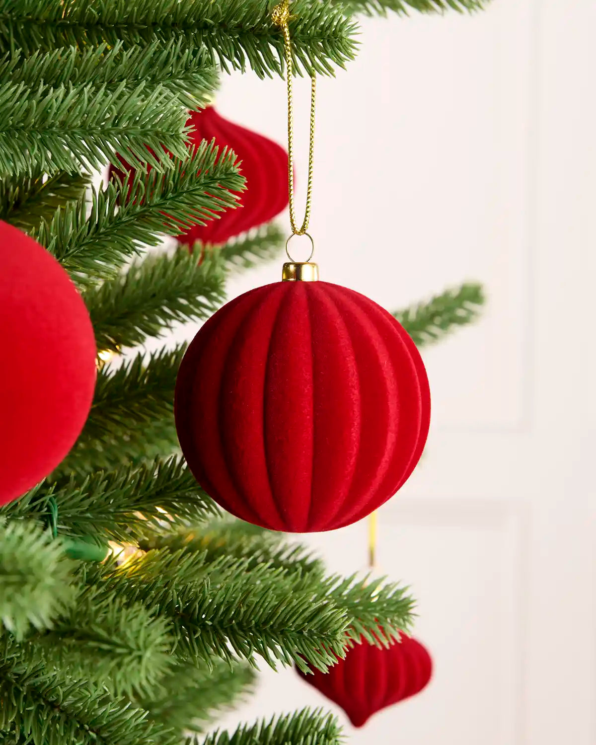 A red ribbed velvet Christmas bauble ornament hanging from a green artificial Christmas tree with a gold hanging string, against a soft white background.
