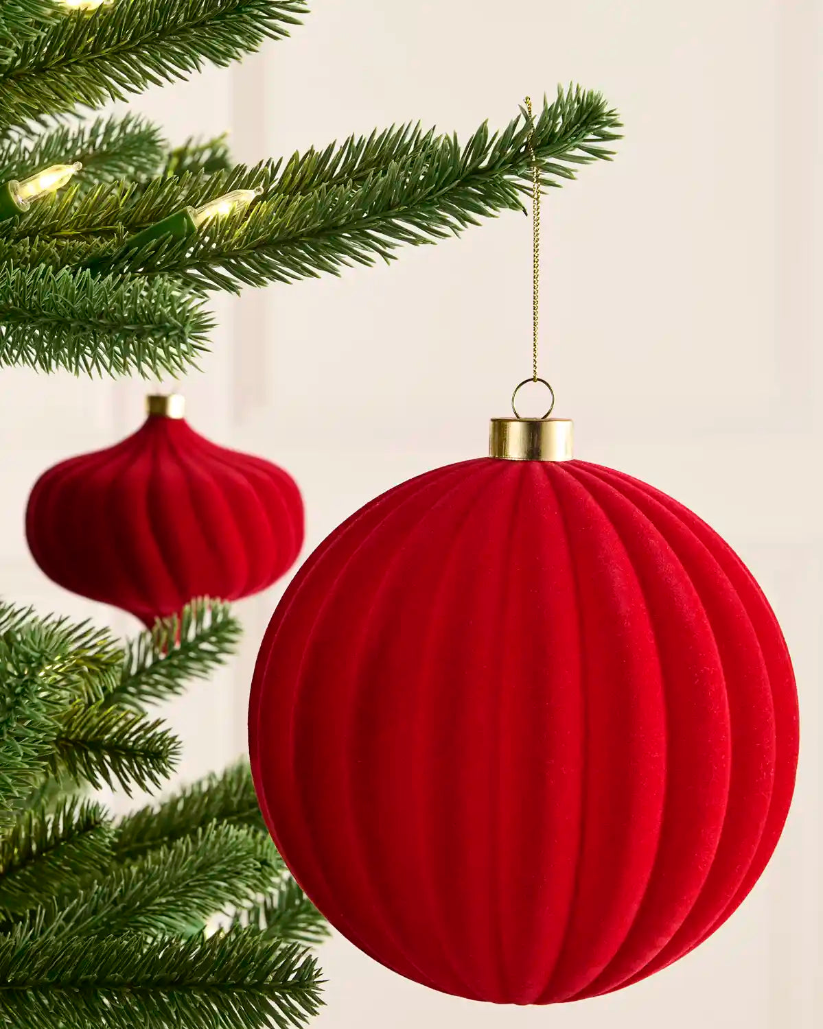 A red ribbed velvet Christmas bauble ornament hanging from a green pine tree branch, with a second matching ornament in the background.