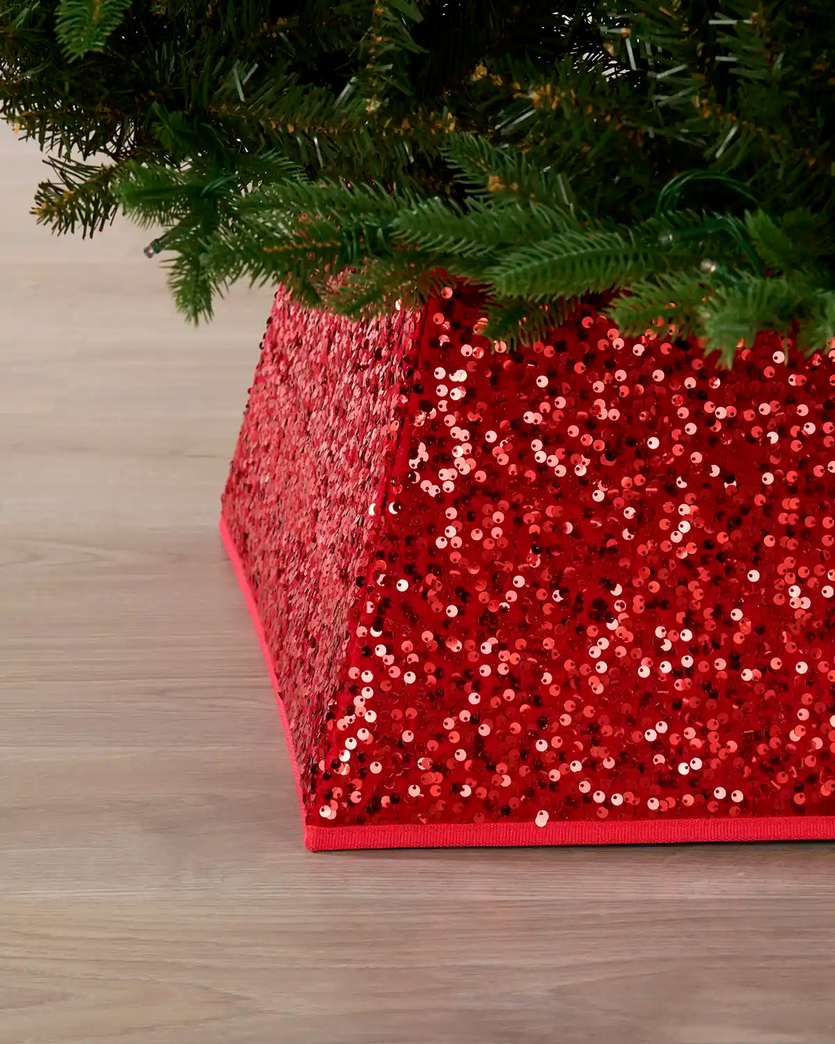 A red sequined tree collar with a geometric tiered design, shown under the branches of a Christmas tree on a light wood floor.