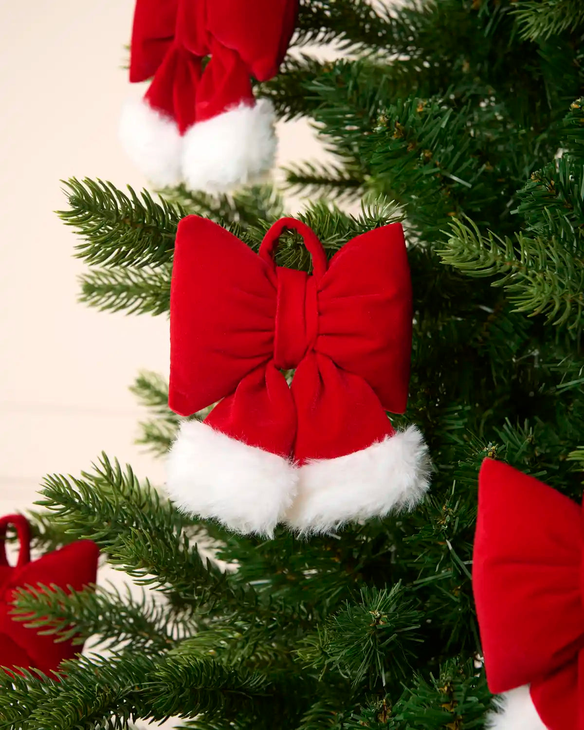 A red velvet Santa hat ornament shaped like a bow with white faux fur trim, hanging on a green Christmas tree.