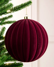 A ribbed, burgundy velvet Christmas bauble ornament hanging from a green pine tree branch against a soft white background.