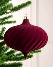 A ribbed, onion-shaped Christmas ornament in burgundy velvet hanging from a green pine tree branch against a soft off-white panelled background.