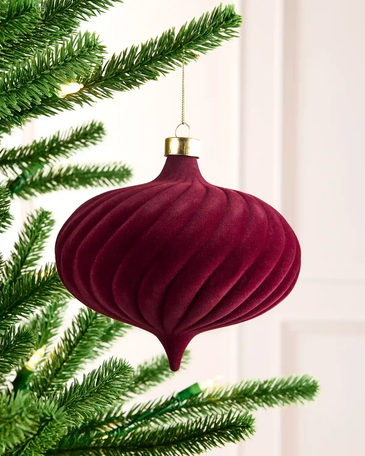 A ribbed, onion-shaped Christmas ornament in deep burgundy velvet with a gold cap and gold hanging string, hanging from a green pine tree branch against a soft white background.