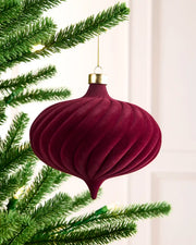 A ribbed, onion-shaped Christmas ornament in deep burgundy velvet with a gold cap and gold hanging string, hanging from a green pine tree branch against a soft white background.