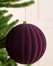 A ribbed, pleated Christmas ornament in  purple velvet, hanging from a gold cap on a gold string next to green pine tree branches.