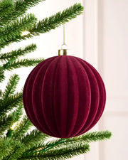 A ribbed, pleated Christmas tree ornament in deep burgundy velvet hanging from a green pine tree branch with warm string lights.