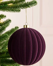 A ribbed, purple velvet Christmas ornament hanging from a green pine tree branch with warm string lights in the background.
