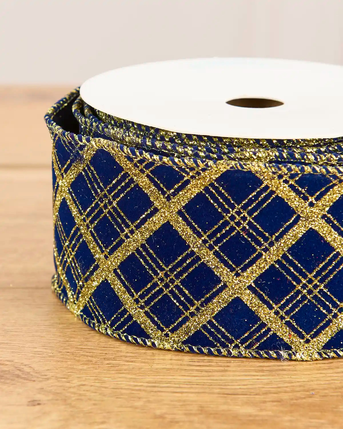 A roll of royal blue velvet ribbon with a glittery gold plaid pattern, wound on a white spool and photographed on a light wood surface.