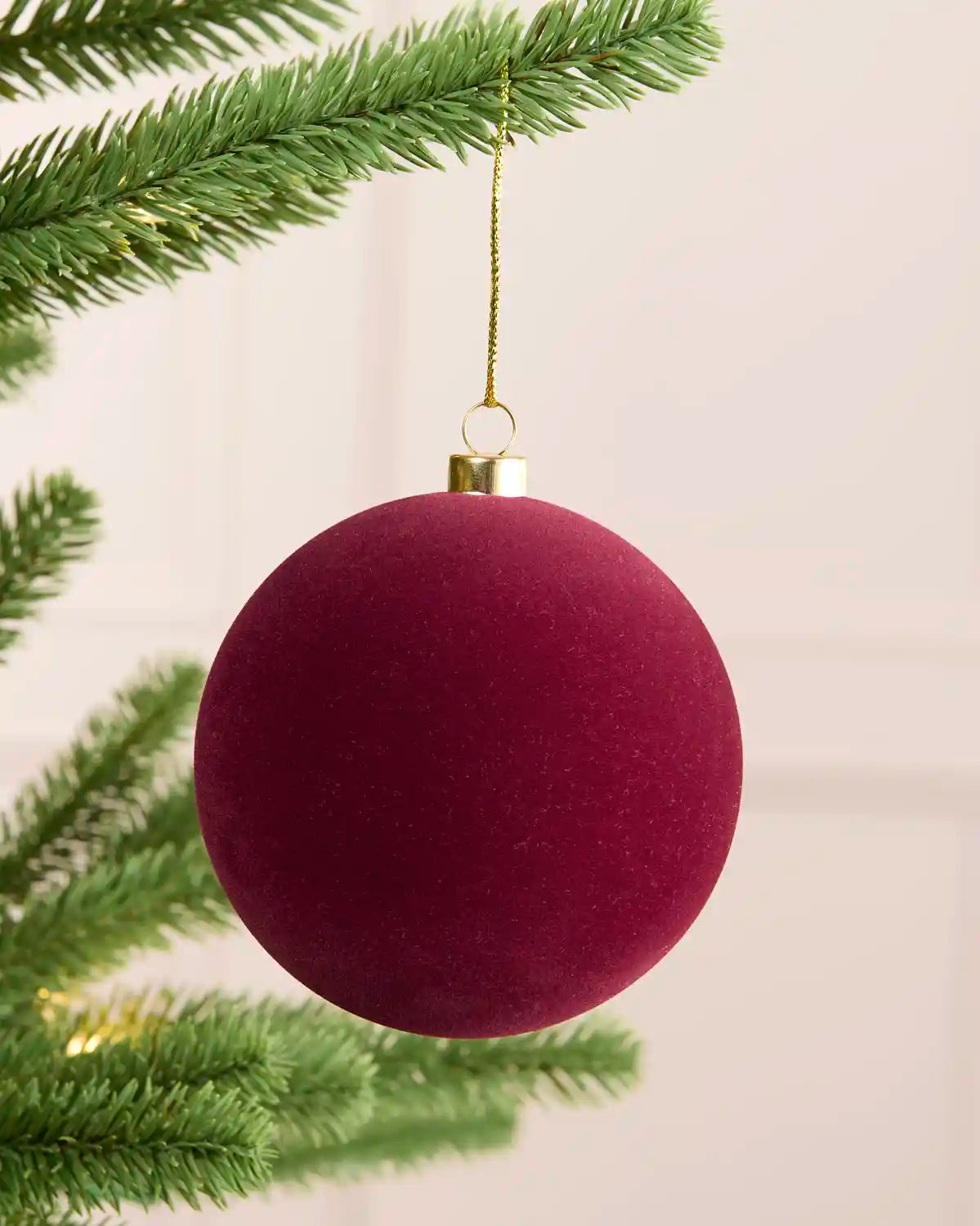 A round, burgundy velvet Christmas ornament hanging from a gold cap and gold string, suspended from a green pine tree branch against a soft, light beige background.