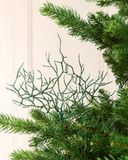 A sage green glittery coral tree topper ornament displayed on the branches of a green artificial Christmas tree against a soft cream-colored paneled background.