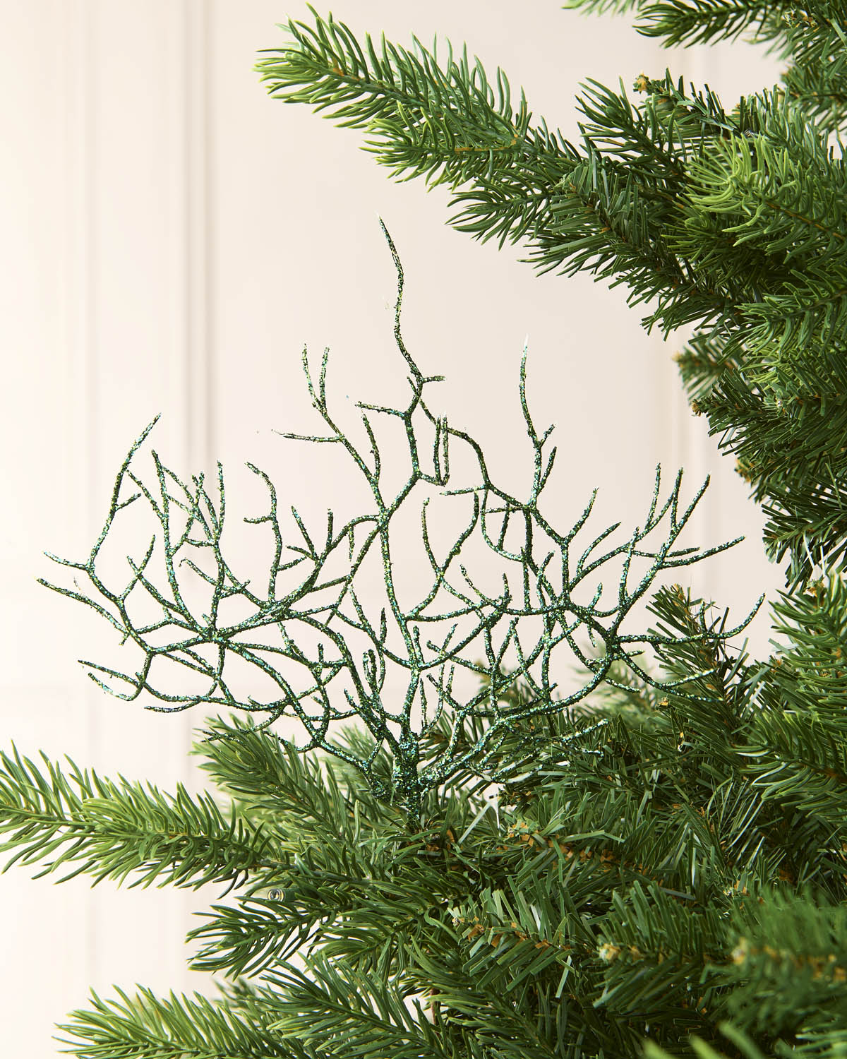 A sage green glittery coral tree topper ornament displayed on the branches of a green artificial Christmas tree against a soft cream-colored paneled background.