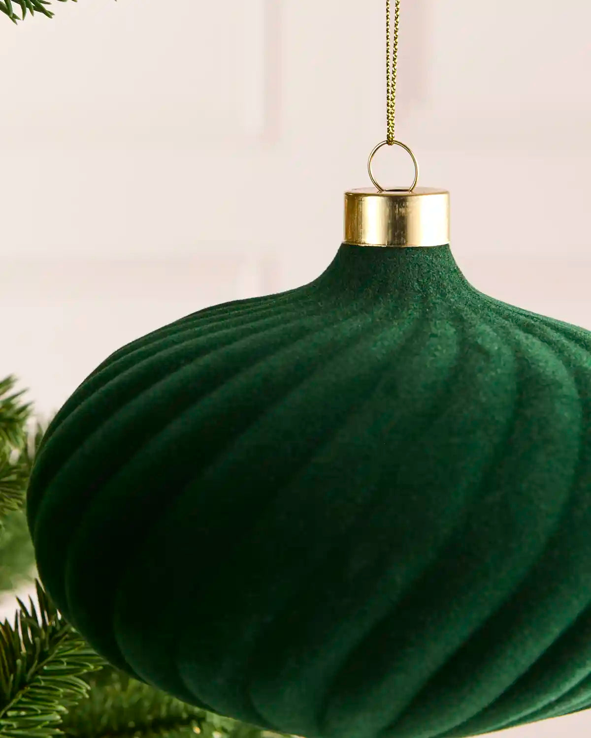 A sage green ribbed Christmas ornament hanging from a gold cap with a gold chain, shown close up against a soft light background with pine branches at the edge.