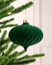 A sage green ribbed velvet Christmas ornament hanging from a gold cap on a gold string, positioned next to green pine tree branches with warm string lights.