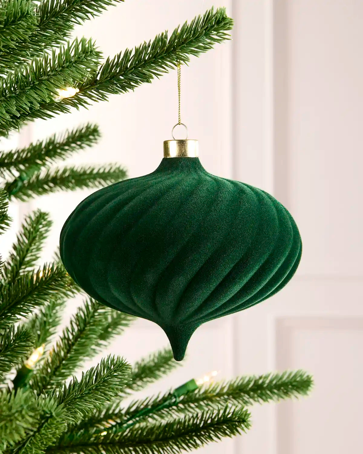 A sage green ribbed velvet Christmas ornament hanging from a gold cap on a gold string, positioned next to green pine tree branches with warm string lights.