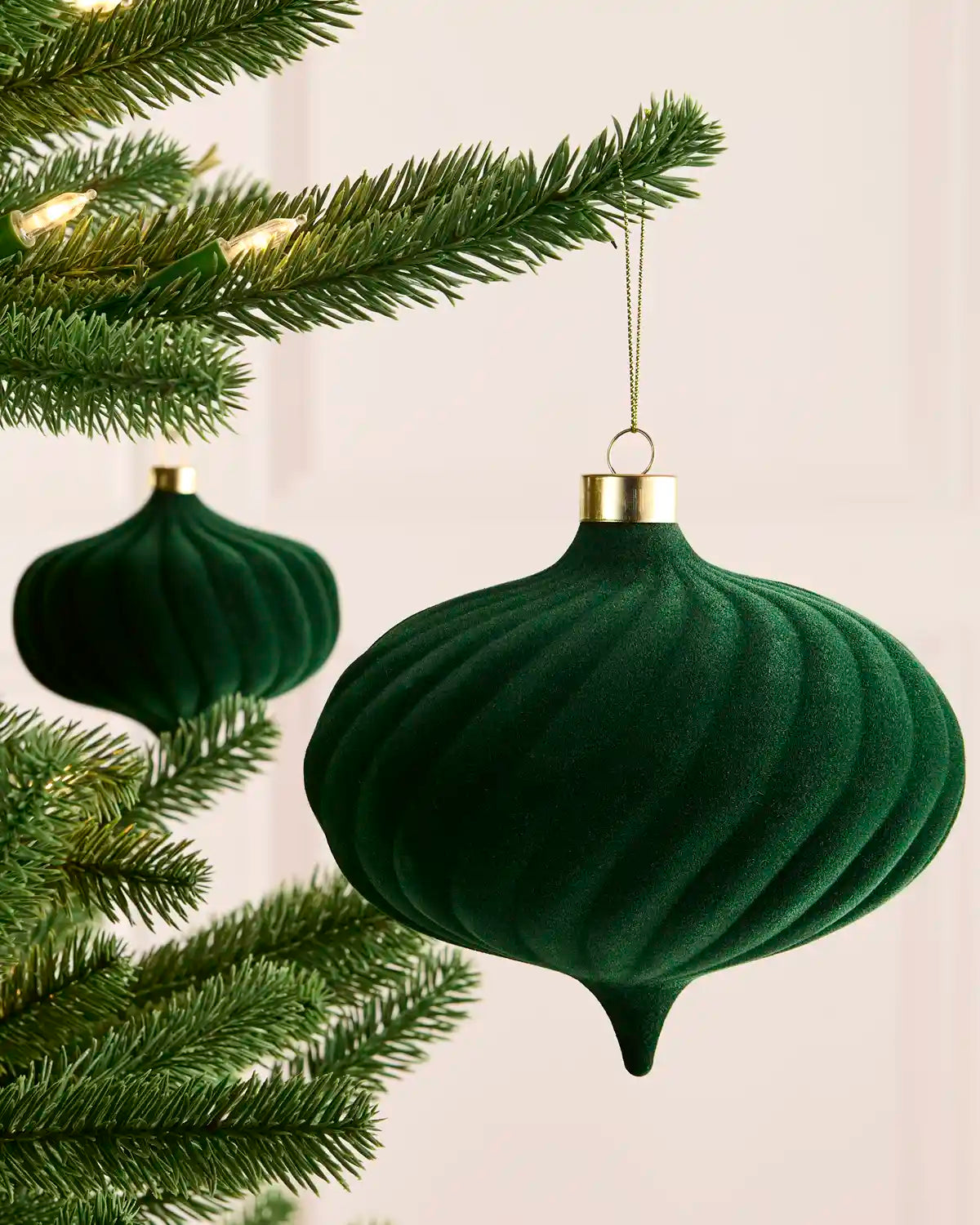 A sage green ribbed velvet Christmas ornament hanging from a gold cap on a tree branch, with a second matching ornament blurred in the background.