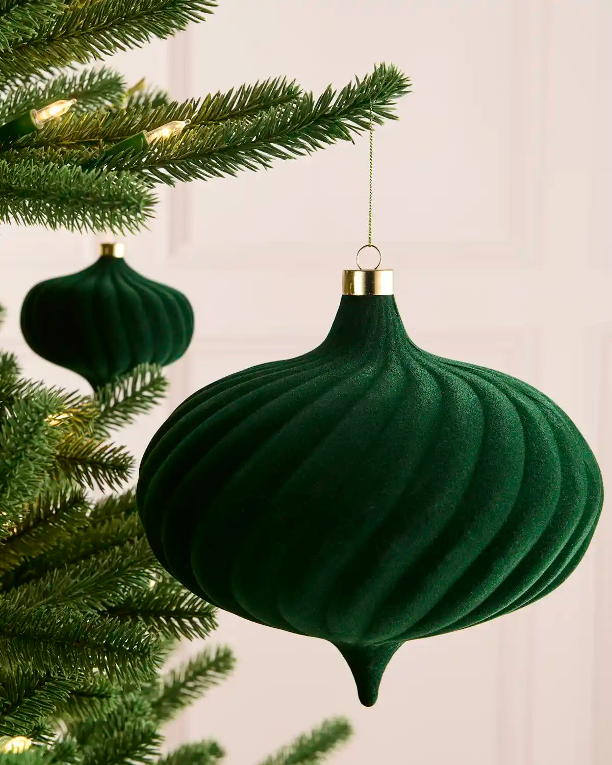 A sage green ribbed velvet Christmas ornament hanging from a gold cap on a tree branch, with another ornament and green tree branches in the background.