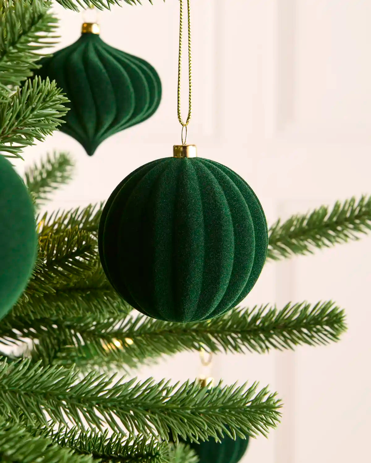 A sage green ribbed velvet Christmas ornament hanging from a gold string on a pine tree branch, with other matching ornaments partially visible in the background.